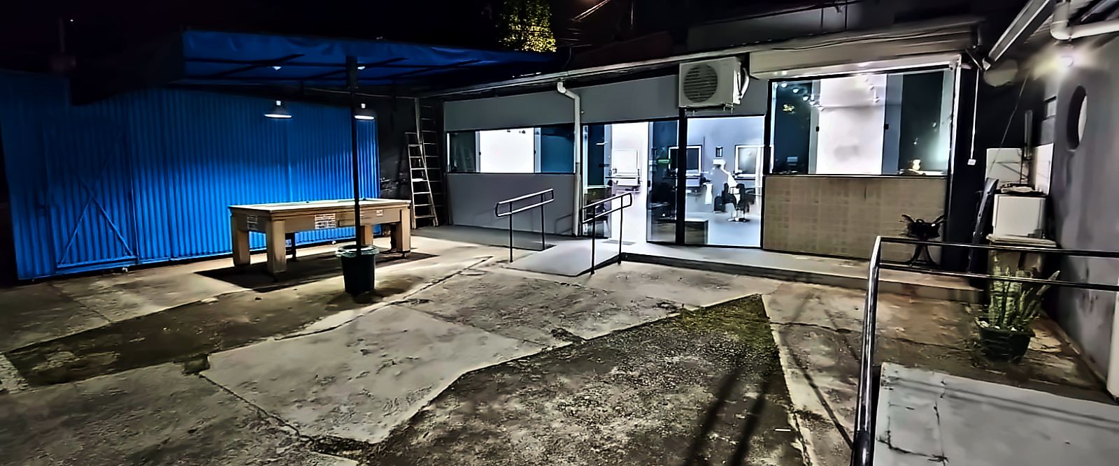 A frente da barbearia à noite, na Rua Hidra: o pátio coberto, a rampa com corrimão e a luz acesa dentro do salão.