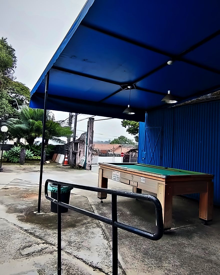O pátio da barbearia de dia, sob a tenda azul, com a rua do bairro ao fundo